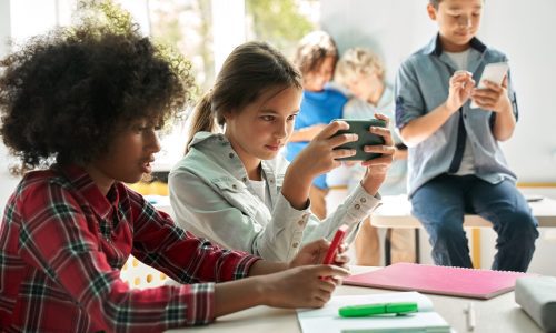 Diverse multiethnic kids students using smartphones in classroom. Multicultural children holding devices having fun with mobile phones apps playing games and checking social media at school.
